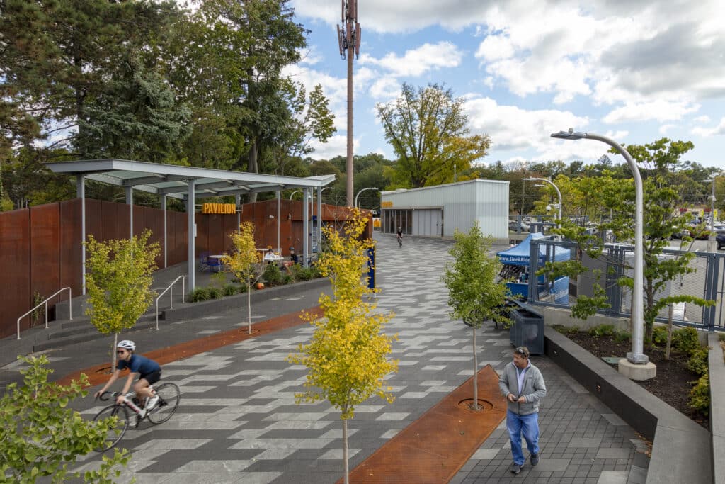 A cyclist rides along a paved, tree-lined walkway near a modern building with a "PATIOS" sign, reminiscent of scenes near the Tappan Zee, while a person walks nearby under a partly cloudy sky.