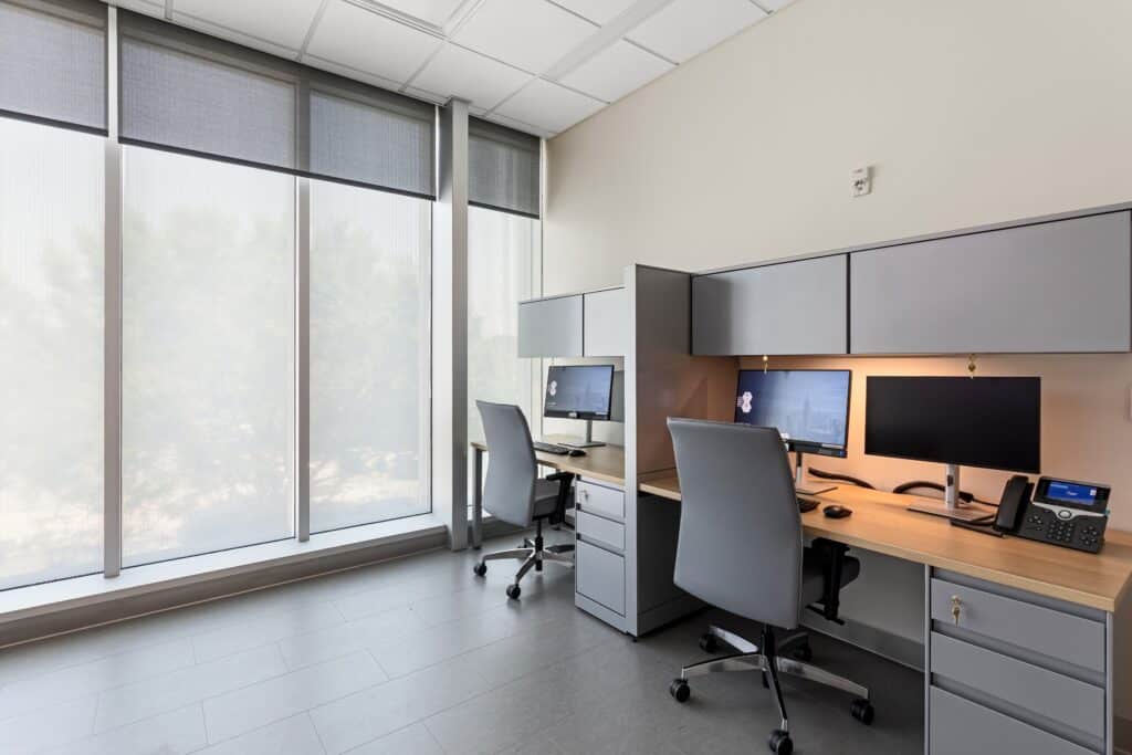 Modern office with two workstations, each with a computer, chair, phone, and overhead cabinets. Large windows on the left let in natural light, creating a bright and inviting environment reminiscent of Patchogue Ambulatory’s commitment to clean and organized workspaces.