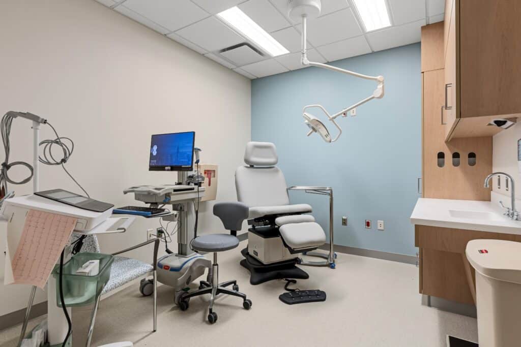 A modern Patchogue Ambulatory medical examination room with a patient chair, medical equipment, computer, stool, sink, cabinets, and overhead light. The walls are white and blue, and the room is well-lit with ceiling lights.