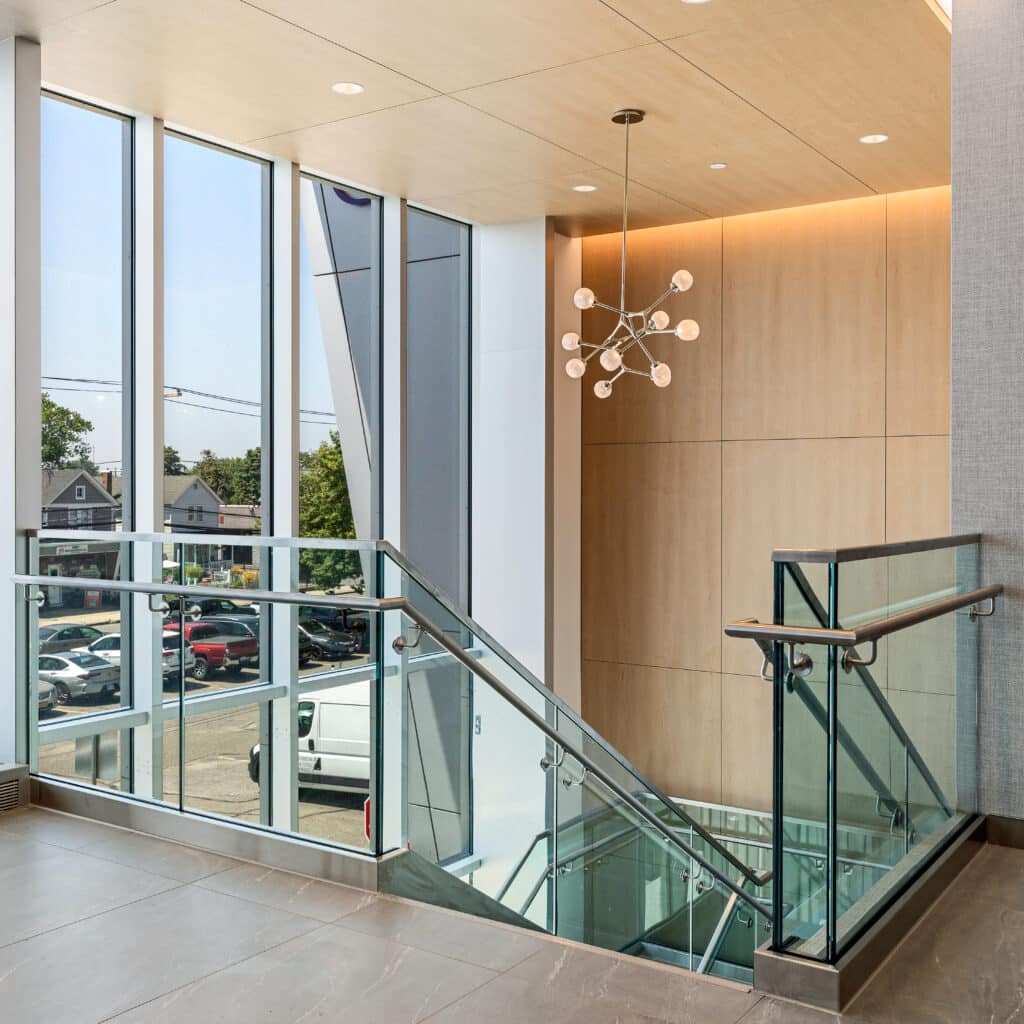 A modern stairwell with glass railings, a geometric light fixture, and large windows that let in natural light creates an inviting atmosphere within Patchogue Ambulatory. From the landing, cars and houses are visible outside, blending exterior views with the center’s contemporary design.