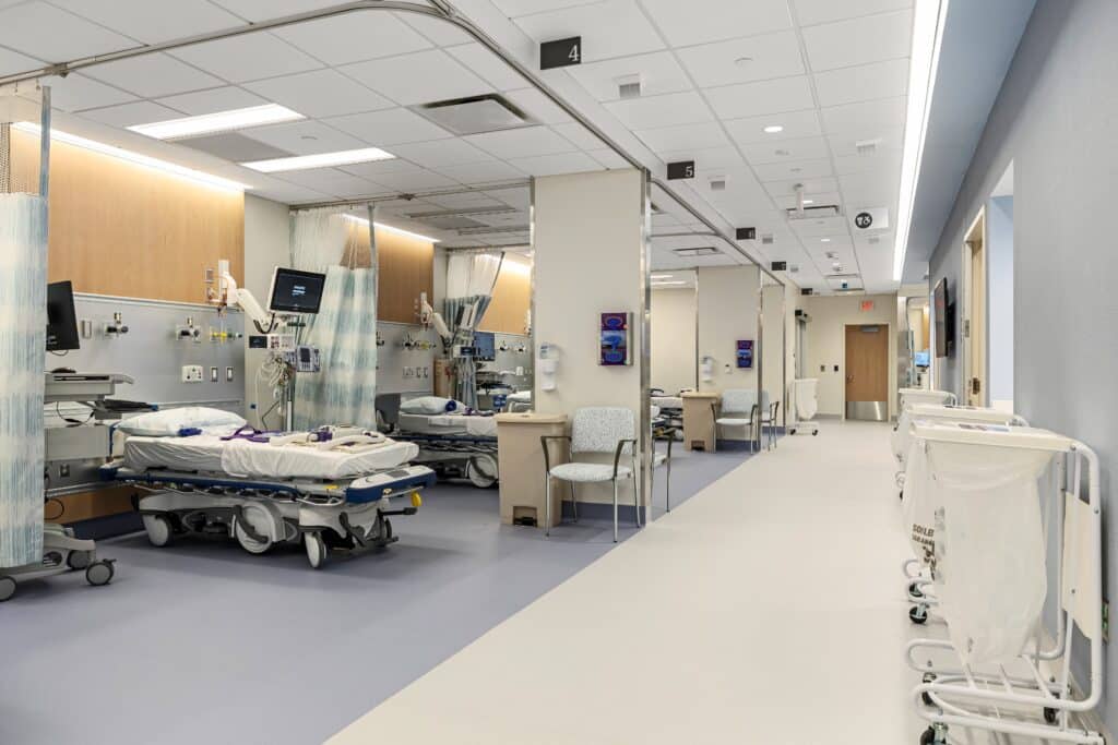 A clean, modern hospital ward at Patchogue Ambulatory features empty patient beds, medical equipment, monitors, chairs, and privacy curtains along a wide, bright hallway. Linen carts are lined up on the right side of the hallway.