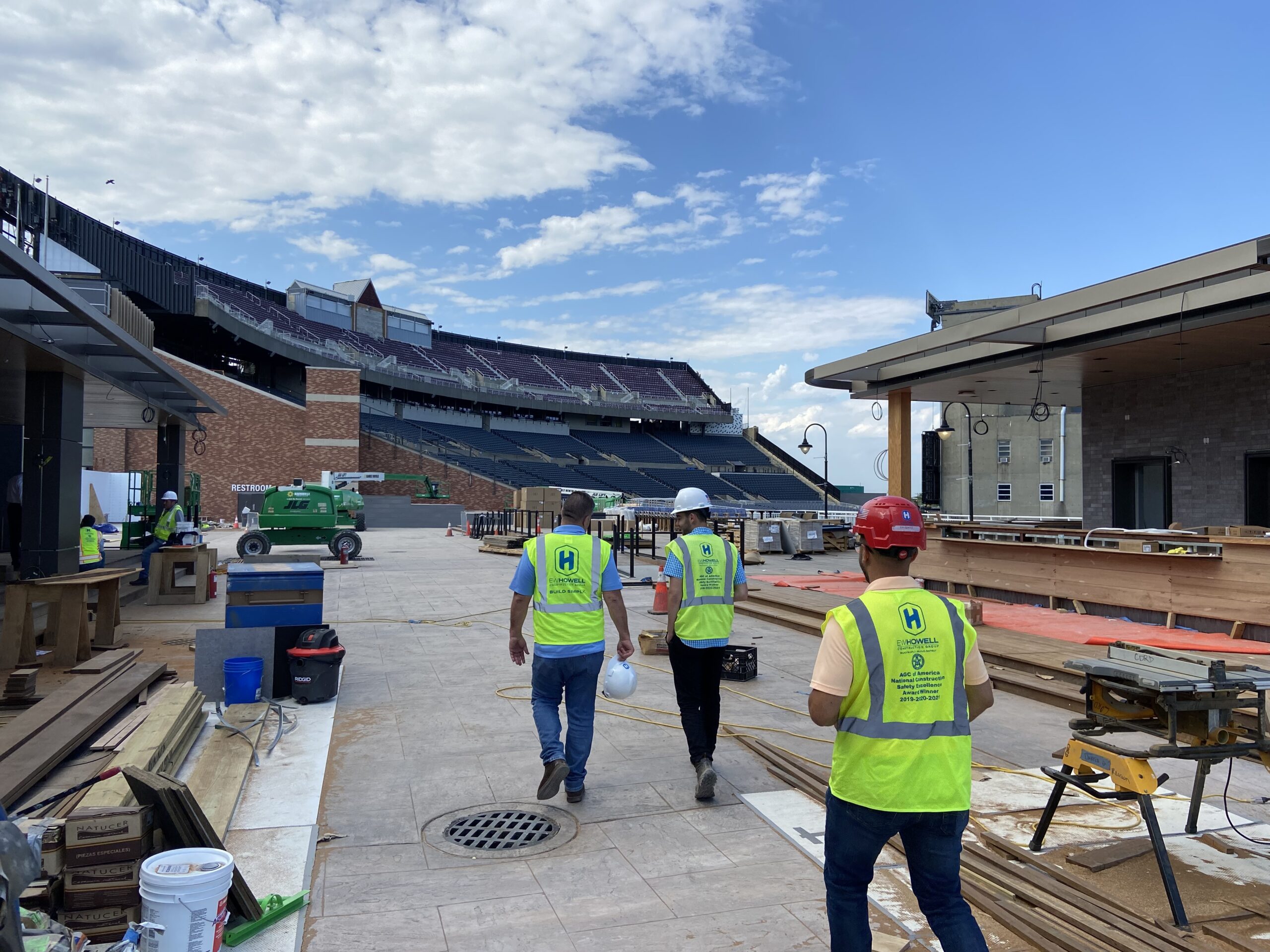 Construction workers in safety vests and helmets walk through an outdoor stadium area under construction, with building materials and equipment scattered around and empty seating visible in the background.