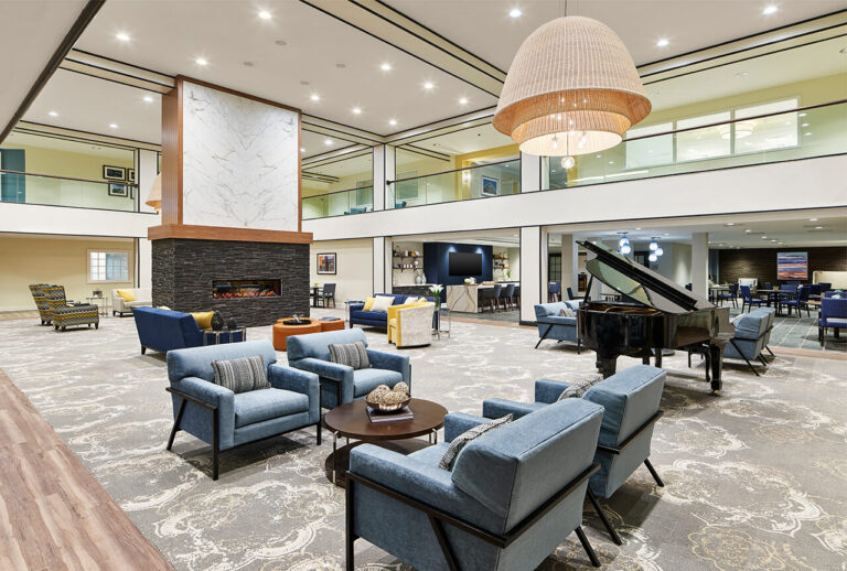 A spacious, modern lounge with blue armchairs, a grand piano, fireplace, and large hanging light fixture. The room features high ceilings, an upstairs balcony, and a bright, inviting atmosphere.