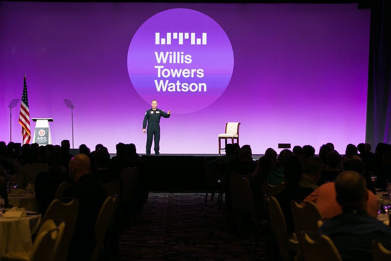 A speaker stands on stage in front of an audience, with a large purple screen behind displaying the Willis Towers Watson logo. An empty chair, the U.S. flag, and a podium are also on the stage.