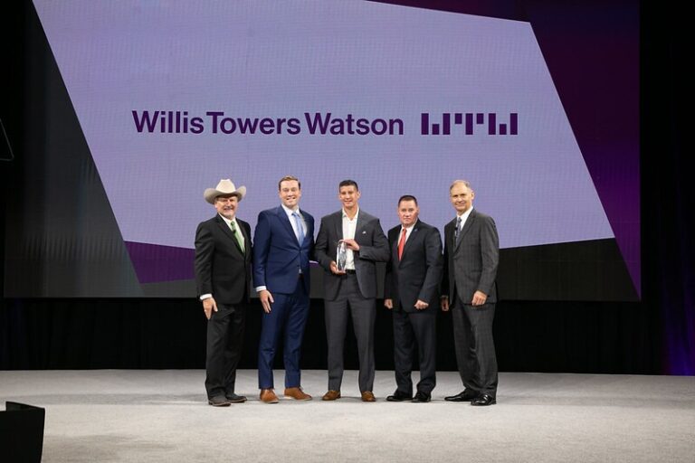 Five men in suits stand on a stage, one holding an award. Behind them is a large screen displaying the Willis Towers Watson logo and name. The background is purple and white.