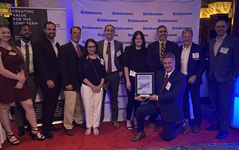A group of twelve people, dressed in business attire, pose together on a red carpet in front of a "Business News" backdrop. One person kneels in front holding a framed 204 Fastest Growing Companies award.