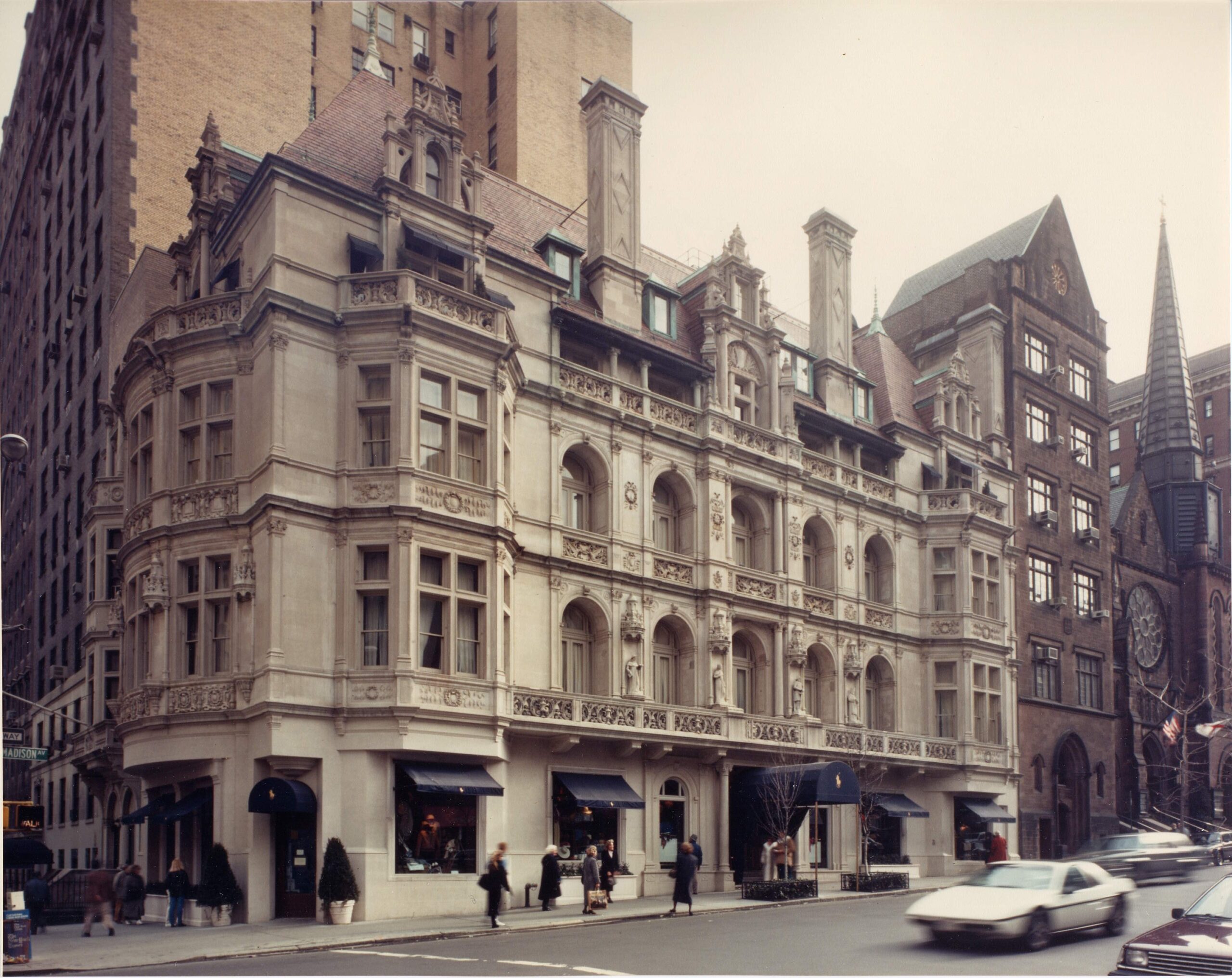 A historic stone building with ornate architectural details and arched windows stands on a city street corner. People walk along the sidewalk and cars drive by. Neighboring buildings and a church with a clock are visible.
