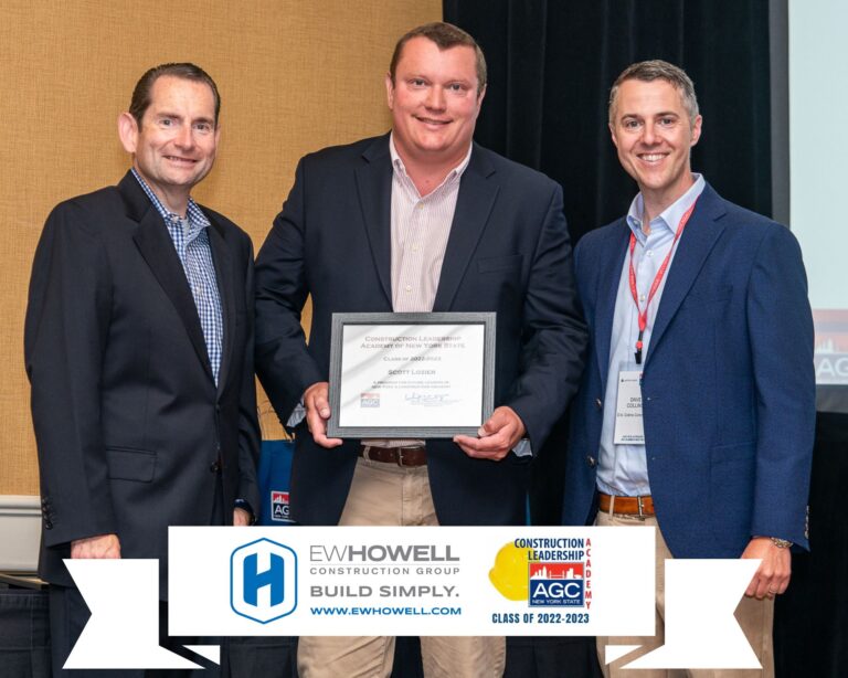 Three men stand together, smiling. The man in the center holds a framed certificate. They are dressed in business attire. In front of them are logos for E.W. Howell Construction Group and AGC, with a banner for “Class of 2022-2023.”.