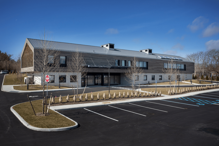 A modern two-story building with large windows, a sloped metal roof, and an adjacent parking lot with marked space, including accessible parking, on a clear day with blue sky. Leafless trees line the front walkway.