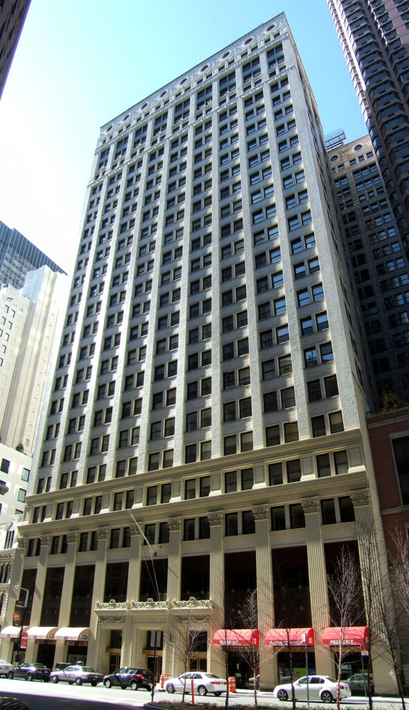 A tall, rectangular office building at 80 Maiden Lane with many windows, a beige facade, and decorative columns at the entrance, surrounded by other high-rise buildings and several parked cars along the street.