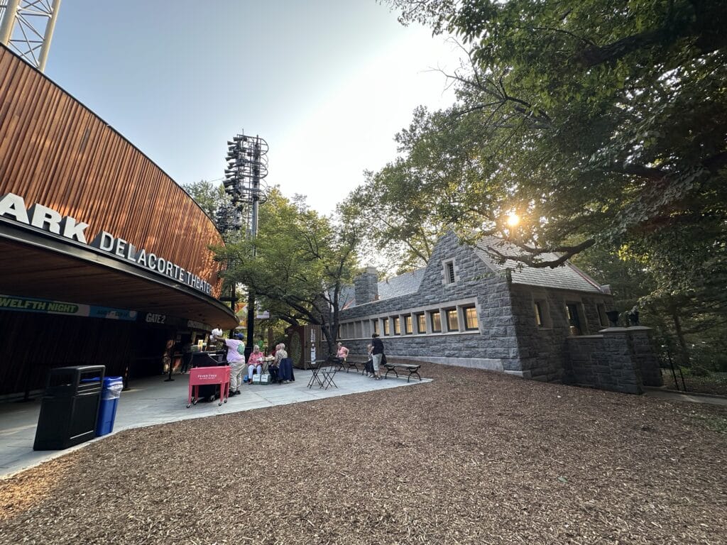 People sit at tables near the Delacorte Theater entrance in a park, framed by a modern wood structure on the left and a stone building on the right, surrounded by trees glowing in the sunset.