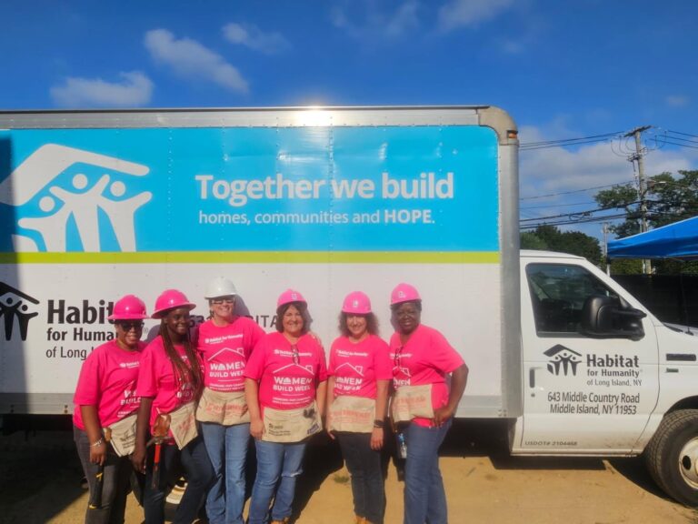Six women in pink shirts and hard hats stand smiling in front of a Habitat for Humanity truck with a sign that reads, “Together we build homes, communities and HOPE.”.